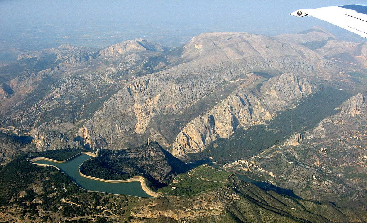 El Chorro from the air: the village and the lower reservoir are at bottom right, the Gaitanes gorge is the cleft left of centre, and the Frontales are the sunlit south-facing walls rising above the pine forest between the two. The upper car park and the track towards the wall lie between the village and the wall.