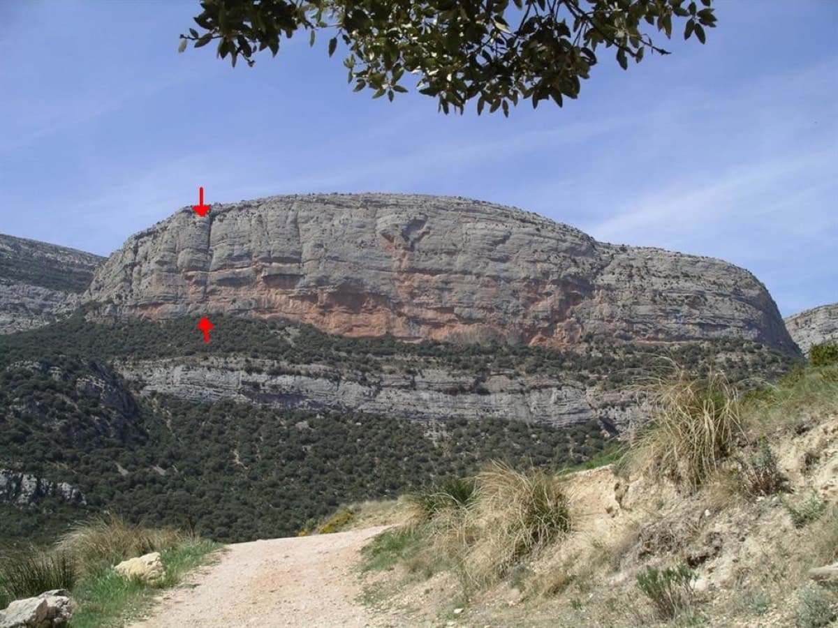 The Roca dels Arcs from the approach track. The two red arrows mark the top and bottom of the Montse Pueyo corner at the left end of the wall; El Somni de Quimfer starts a few metres right of it.
