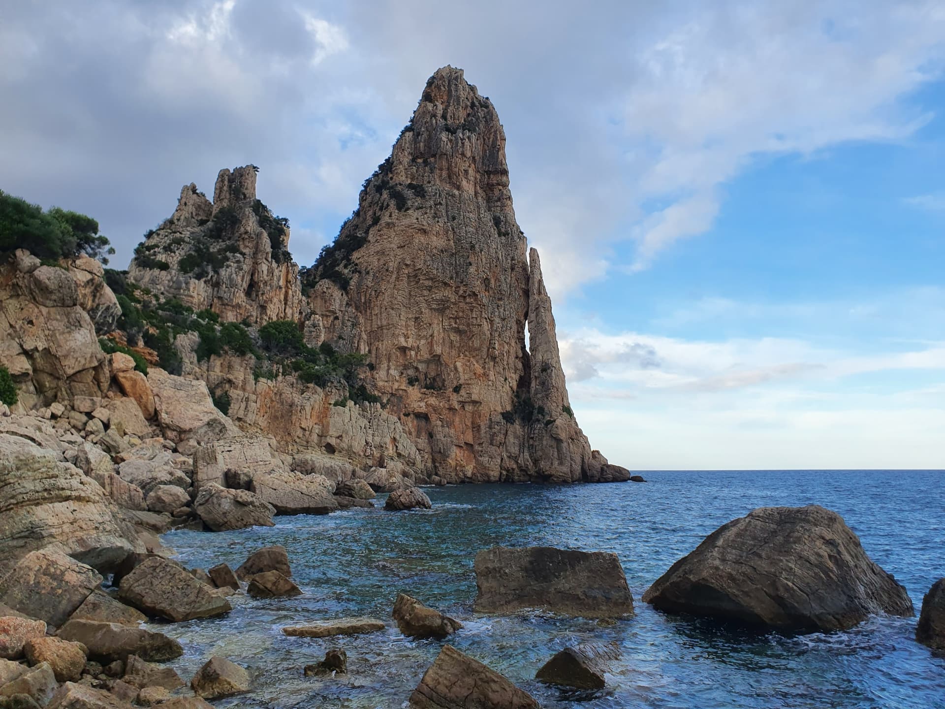 Pedra Longa, the 128 m limestone tower on the Baunei coast, seen from the shoreline rocks at its foot.