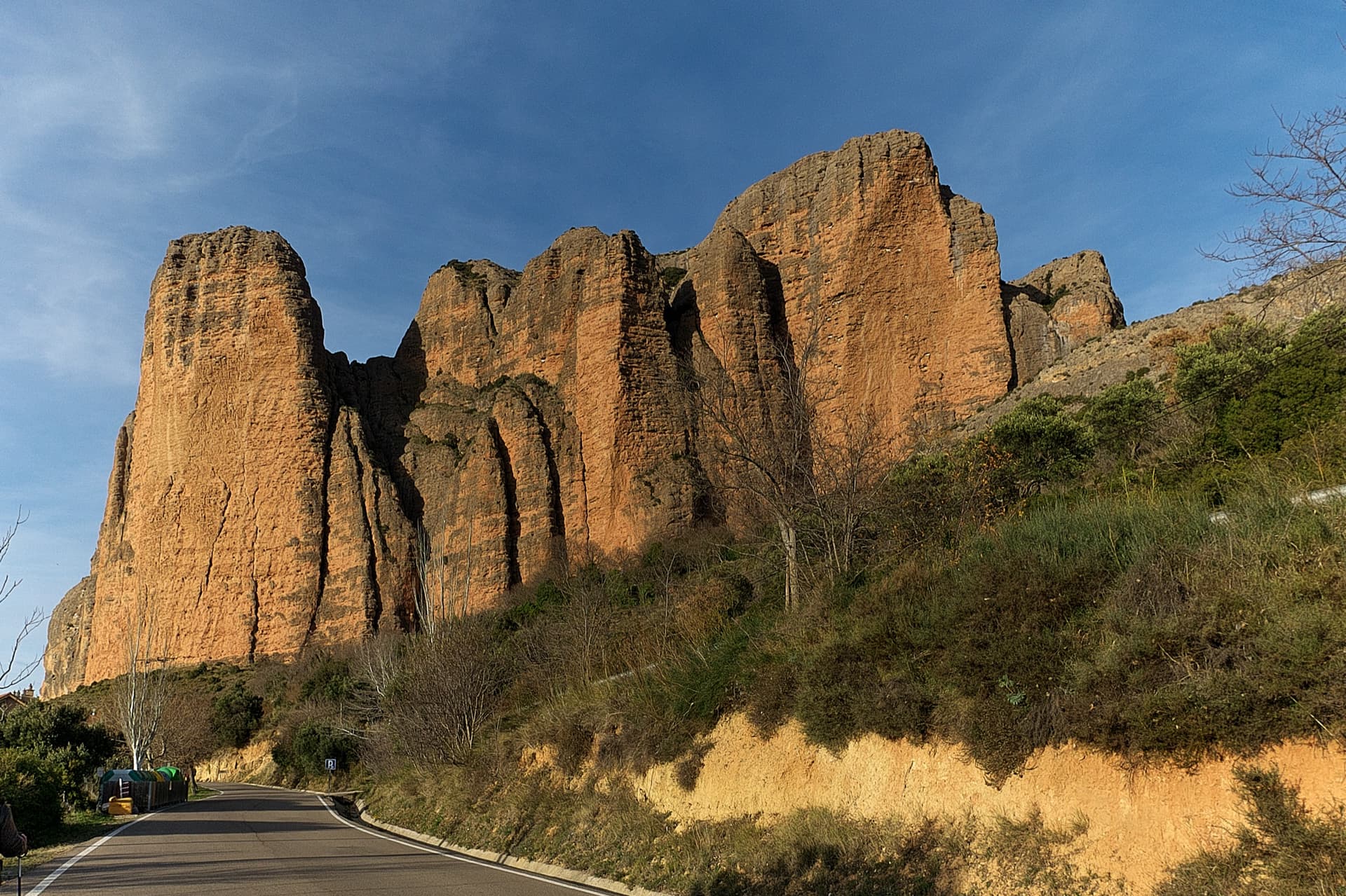 The south faces of the main Riglos wall in low sun, seen from the road into the village: Mallo Pisón on the left and the wall running right towards Mallo Frechín and La Visera at the right-hand end.