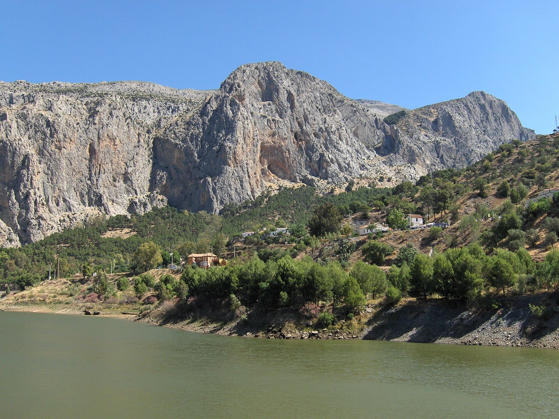 The Frontales cliffs above El Chorro seen across the Tajo de la Encantada reservoir, with the big Poema de Roca cave at the foot of the central dome. El Corazón que no Siente is on this wall; the line is not marked.